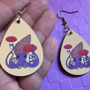 May include: Two pairs of yellow teardrop shaped earrings with a purple octopus wearing a brown snail shell and two red mushrooms on its head.