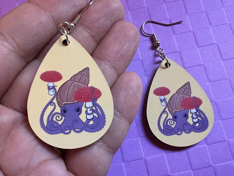 May include: Two pairs of yellow teardrop shaped earrings with a purple octopus wearing a brown snail shell and two red mushrooms on its head.