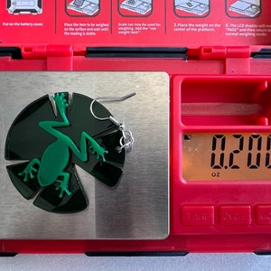 May include: A green frog earring with a silver hook, weighing 0.200 ounces on a digital scale.
