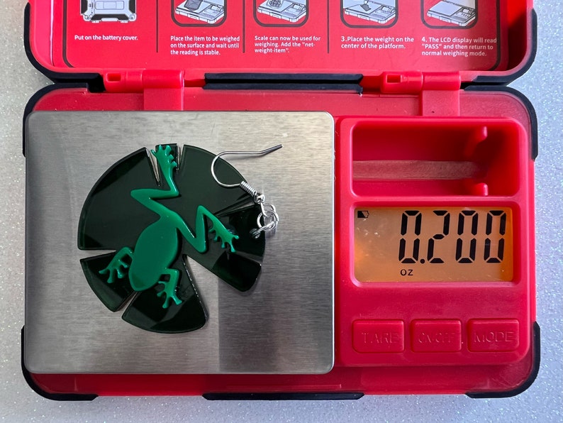May include: A green frog earring with a silver hook, weighing 0.200 ounces on a digital scale.