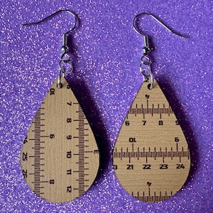 May include: A pair of wooden teardrop-shaped earrings with ruler markings. The earrings are made of light brown wood and have silver hooks.