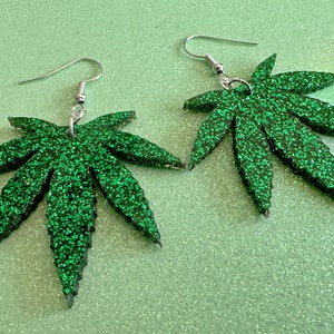 May include: A pair of green glitter marijuana leaf earrings. The earrings are made of resin and have a silver hook.