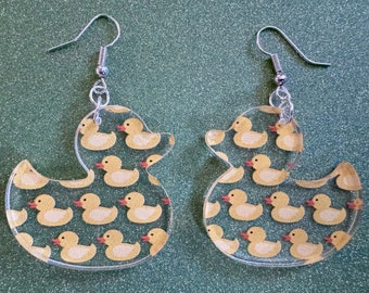 May include: A pair of clear acrylic earrings shaped like rubber ducks. The ducks are decorated with a pattern of smaller yellow rubber ducks.