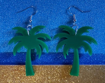 May include: A pair of green palm tree earrings with silver hooks. The earrings are on a blue and gold sparkly background.