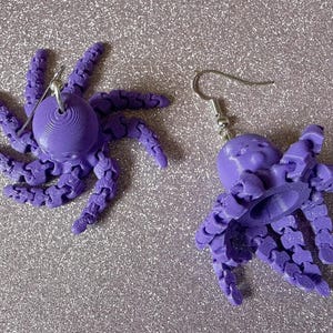 May include: A pair of purple octopus earrings. Each earring features a detailed octopus design with articulated tentacles and a round head. The earrings are suspended from silver-colored ear wires.