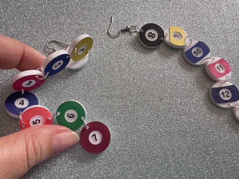 May include: A set of 12 colorful, round, numbered earrings, each with a white background and black numbers. The earrings are connected by silver jump rings and are in the shape of a circle.