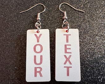 May include: A pair of white rectangular earrings with silver hooks. The earrings have the words "YOUR" and "TEXT" printed vertically in pink.