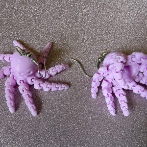 May include: A pair of lavender octopus earrings. Each earring features a detailed octopus design with multiple tentacles and a silver-colored ear wire. The earrings are set against a glittery, light purple background.