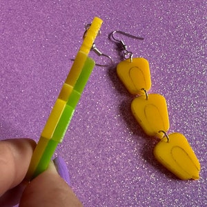 May include: A pair of earrings with a yellow and green geometric design on one earring and a chain of three yellow teardrop shapes on the other earring.