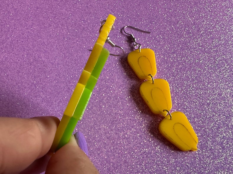 May include: A pair of earrings with a yellow and green geometric design on one earring and a chain of three yellow teardrop shapes on the other earring.