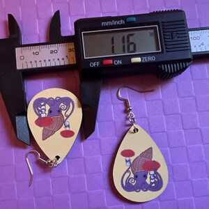 May include: Two wooden earrings with a yellow background and a purple octopus holding a red mushroom and a brown shell. The earrings are measured with a caliper that reads '1.16' in the display. The earrings are approximately 1.16 inches in length.