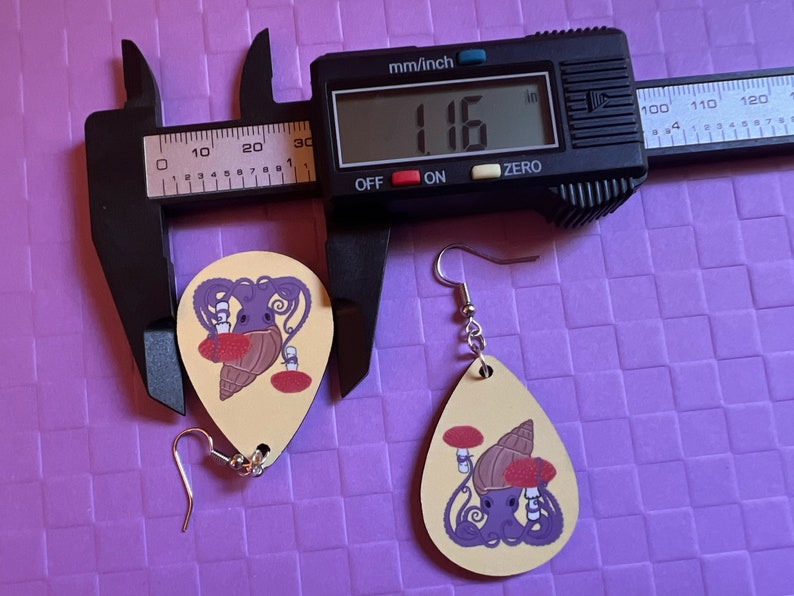 May include: Two wooden earrings with a yellow background and a purple octopus holding a red mushroom and a brown shell. The earrings are measured with a caliper that reads '1.16' in the display. The earrings are approximately 1.16 inches in length.