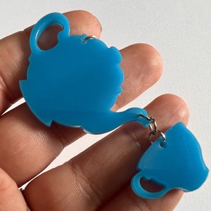 May include: A teal blue teapot and teacup charm with a silver chain connecting them.
