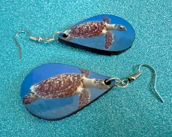 May include: A pair of teardrop-shaped earrings featuring a sea turtle design. The earrings have a blue background with a detailed image of a turtle. The earrings are made of a lightweight material and have silver-colored ear wires.