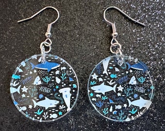 Pode incluir: Um par de brincos de argola de prata com pingentes redondos de acrílico transparente. Os pingentes têm um padrão azul e branco de tubarões, estrelas do mar e algas marinhas.