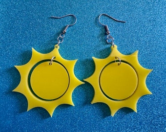 May include: A pair of yellow sun-shaped earrings with a silver hook closure. The earrings are made of acrylic and have a shiny finish.