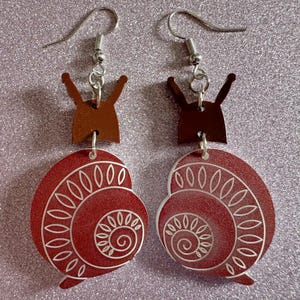 May include: A pair of red snail earrings with silver hooks. The snail shells have a white swirl design and are accented with brown details.