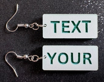 May include: A pair of silver earrings with a white rectangular charm. The charm has the words "TEXT" and "YOUR" written in green.