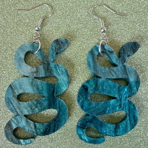 May include: A pair of teal blue snake-shaped earrings with silver hooks. The earrings are made of a marbled material with a glittery finish.