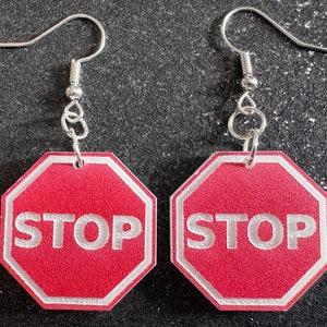 Puede incluir: Un par de pendientes de señal de stop roja con ganchos plateados. Las señales de stop son octogonales y tienen letras blancas que dicen "STOP".