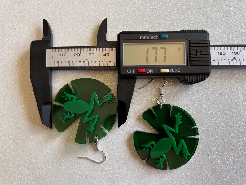 May include: Green frog earrings with a lily pad design. The earrings are 1.77 inches in diameter.