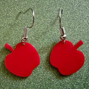May include: A pair of red apple-shaped earrings. Each earring has a bright red, glossy apple pendant with a small stem, hanging from a silver-coloured fish hook ear wire. The earrings are set against a sparkly green background.