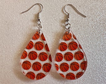 May include: A pair of teardrop-shaped earrings with a basketball pattern. The earrings are made of clear acrylic and have silver hooks.