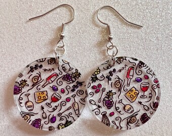 May include: Clear, round earrings with a wine theme. Each earring features a repeating pattern of wine bottles, grapes, cheese, and glasses in black, red, and purple. The word "Vine" is also visible on each earring.