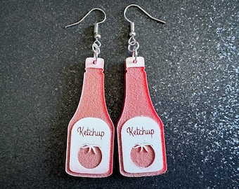 May include: A pair of red ketchup bottle earrings with white accents. The bottles have the word "Ketchup" printed on them and a white tomato image.