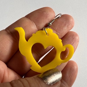 May include: Yellow teapot shaped earrings with a heart cut out in the center. The earrings are made of acrylic and have silver hooks.