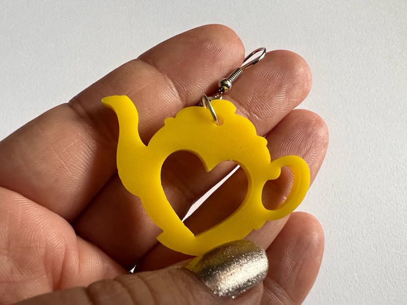 May include: Yellow teapot shaped earrings with a heart cut out in the center. The earrings are made of acrylic and have silver hooks.