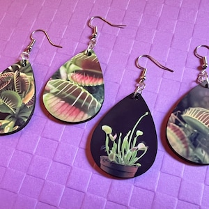 May include: Three pairs of dangle earrings with a teardrop-shaped design. Each earring features a different image of a carnivorous plant. One pair features a close-up of a Venus flytrap, another pair features a close-up of a pitcher plant, and the third pair features a potted sundew plant.