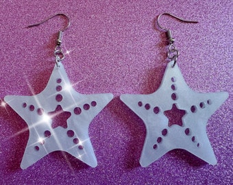 May include: A pair of frosted white star-shaped earrings with a smaller star cut out in the center. The earrings have silver hooks.