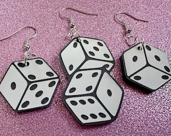 May include: A pair of dangle earrings featuring white dice with black dots. The dice are stacked on top of each other and have a silver hook.