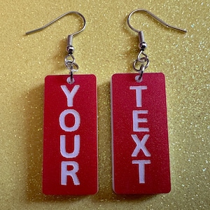May include: A pair of red rectangular earrings with silver hooks. The earrings have white text that reads "YOUR TEXT" on them.