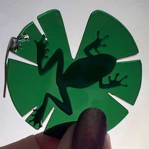 May include: Green acrylic charm featuring a silhouette of a frog on a leaf. The charm has a silver loop for attaching to a necklace or bracelet.