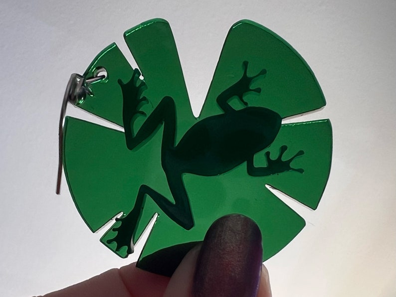 May include: Green acrylic charm featuring a silhouette of a frog on a leaf. The charm has a silver loop for attaching to a necklace or bracelet.