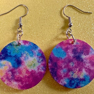 May include: A pair of round tie-dye earrings with silver hooks. The earrings are a mix of pink, blue, and yellow colors.
