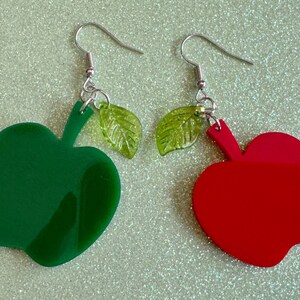 May include: A pair of dangle earrings featuring green and red apple shapes with green leaf accents.