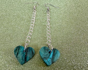 May include: A pair of dangle earrings with a teal and black marbled heart charm on each earring. The earrings are attached to a silver chain.