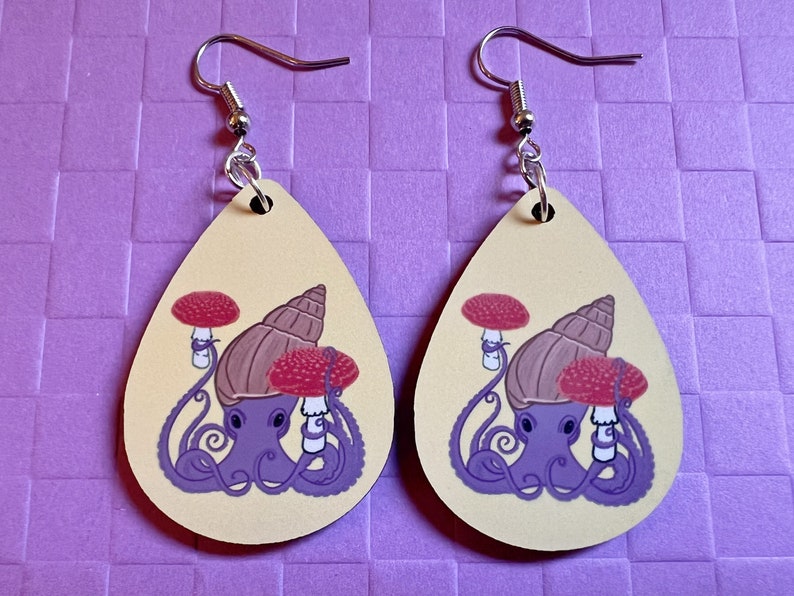 May include: Two teardrop-shaped earrings with a light yellow background. Each earring features a purple octopus with a brown snail shell on its head. The octopus is holding two red and white mushrooms. The earrings have silver hooks.