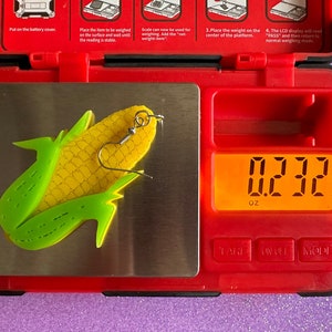 May include: A yellow and green corn on the cob earring with a silver hook, weighing 0.232 ounces on a digital scale.