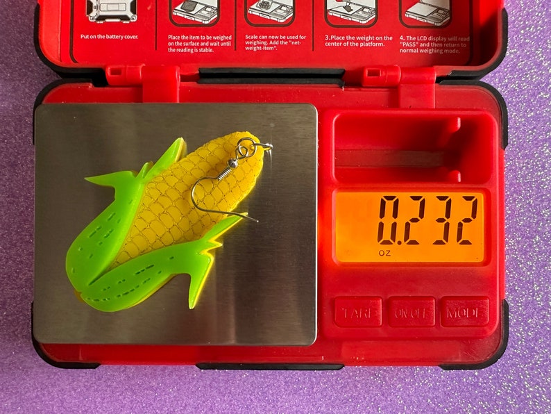 May include: A yellow and green corn on the cob earring with a silver hook, weighing 0.232 ounces on a digital scale.