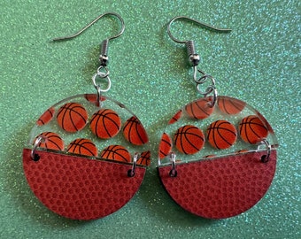 May include: A pair of basketball-themed earrings. Each earring is a clear acrylic circle with a basketball pattern on the top half and a brown textured basketball pattern on the bottom half.