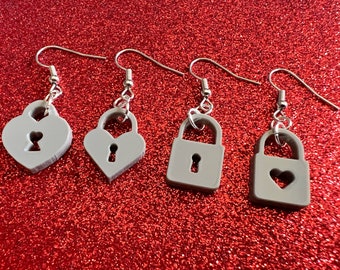 May include: Four pairs of gray earrings with silver hooks. The earrings are shaped like padlocks and hearts, with keyhole cutouts. The earrings are displayed on a red glittery surface.