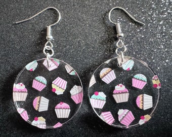 May include: A pair of silver hoop earrings with a clear acrylic charm featuring a repeating pattern of colorful cupcakes.