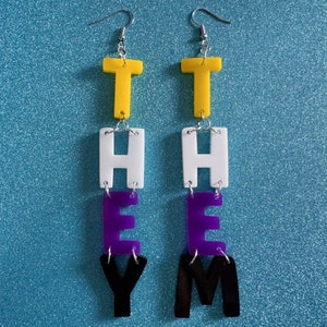 Puede incluir: Un par de pendientes colgantes con la palabra "THEY" escrita en letras acrílicas amarillas, blancas, moradas y negras.