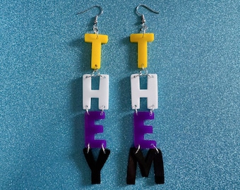 May include: A pair of dangle earrings with the word "THEY" spelled out in yellow, white, purple, and black acrylic letters.