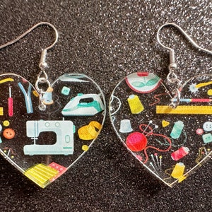 May include: A pair of heart-shaped earrings with a sewing theme. The earrings are made of clear resin and feature a variety of sewing-related images, including a sewing machine, thread, needles, and buttons. The earrings are silver in color and have a hook closure.