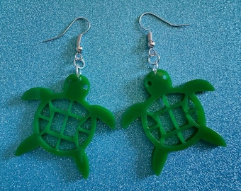 May include: A pair of green turtle earrings with a cutout design. The earrings are made of a lightweight material and have silver hooks.